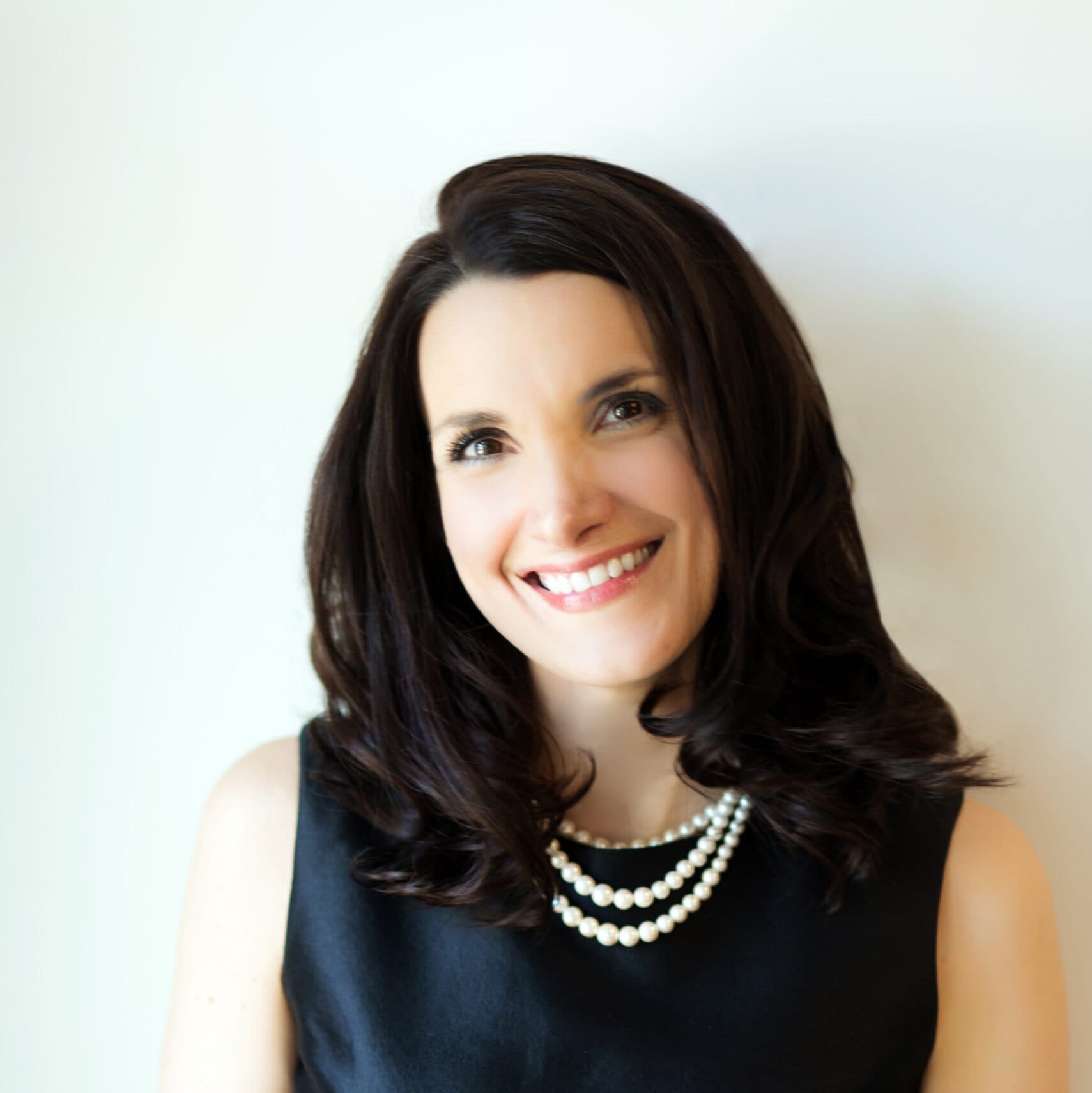 a white woman with dark hair in a black dress and pearls smiles while standing in front of a white wall